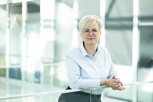 Doc. dr. Jurgita Stravinskienė, MVG centro vadovė, stovi moderniame universiteto interjere, vilkinti šviesius marškinius, žvelgia į kamerą šiltai ir profesionaliai.