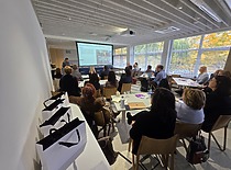 Auditorijoje matosi daug žmonių, sėdinčių ir klausiančiųsi mokymų; priekyje stovi lektorius, rodantis skaidres ar pristatymą